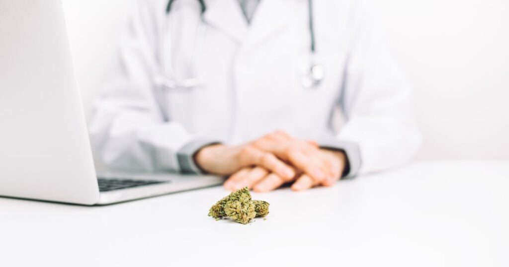 A medical professional in a white lab coat with a stethoscope sits at a desk, hands clasped in front of them. On the white desk surface, a small pile of dried cannabis buds is placed near an open silver laptop, signifying marijuana rescheduling discussions in a clinical setting. The background is clean and bright, emphasizing professionalism
