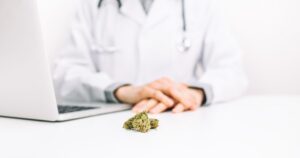 A medical professional in a white lab coat with a stethoscope sits at a desk, hands clasped in front of them. On the white desk surface, a small pile of dried cannabis buds is placed near an open silver laptop, signifying marijuana rescheduling discussions in a clinical setting. The background is clean and bright, emphasizing professionalism
