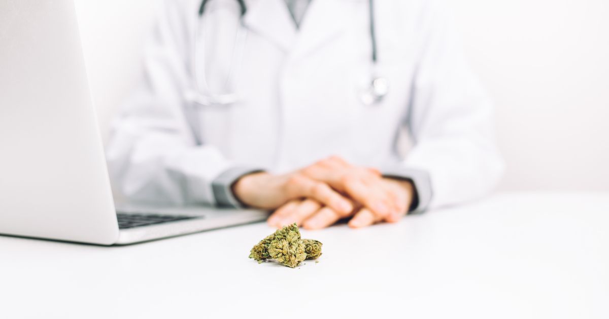 A medical professional in a white lab coat with a stethoscope sits at a desk, hands clasped in front of them. On the white desk surface, a small pile of dried cannabis buds is placed near an open silver laptop, signifying marijuana rescheduling discussions in a clinical setting. The background is clean and bright, emphasizing professionalism