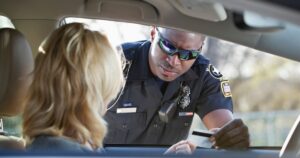 Scene of a traffic stop illustrating the context of Georgia House Bill 496. A police officer in a dark uniform with a 'SWAT' name tag and reflective sunglasses leans into a car window, accepting a white card from the driver. The driver, seen from behind with blonde wavy hair, extends the card from the left side of the frame. The background is blurred, showing hints of greenery and daylight, emphasizing the interaction between law enforcement and the driver