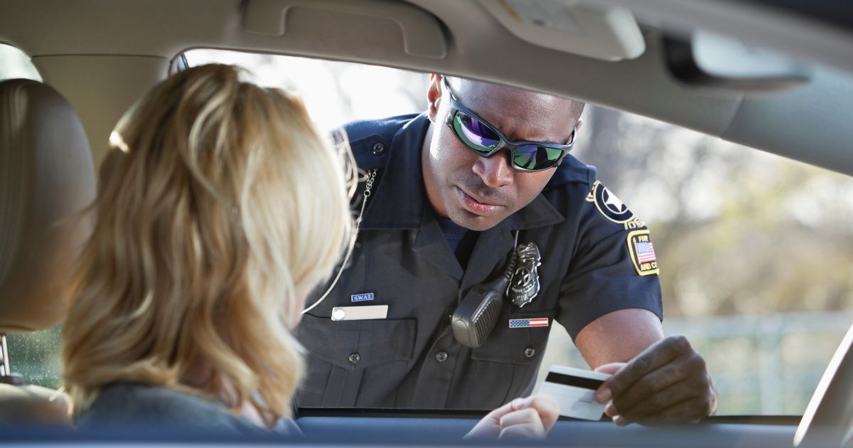 Scene of a traffic stop illustrating the context of Georgia House Bill 496. A police officer in a dark uniform with a 'SWAT' name tag and reflective sunglasses leans into a car window, accepting a white card from the driver. The driver, seen from behind with blonde wavy hair, extends the card from the left side of the frame. The background is blurred, showing hints of greenery and daylight, emphasizing the interaction between law enforcement and the driver