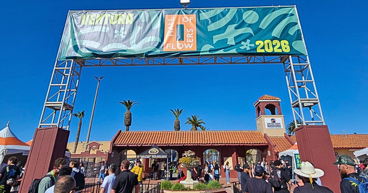 Outdoor scene at the Ventura County Fairgrounds during the Hall of Flowers 2026 event. A large banner with 'HALL OF FLOWERS' and '2026' spans a metal truss above a crowd of attendees. The backdrop features Spanish Colonial Revival architecture, palm trees, and a clear blue sky. Keywords: Hall of Flowers Ventura 2026 recap