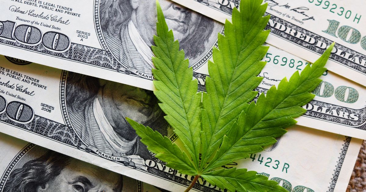 Overhead view of a vibrant green cannabis leaf placed on top of several fanned-out US one hundred-dollar bills. The bills display details like 'Series 2003 A' and 'Series 2006,' with parts of Benjamin Franklin's portrait visible. The textured background adds depth, while the composition highlights the financial connection to cannabis. Keywords: Los Angeles cannabis tax