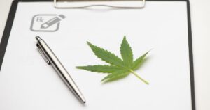 Close-up of a black clipboard holding a white sheet of paper with a green cannabis leaf on the right side. A silver pen lies diagonally across the paper, and a gray 'Rx' icon with a pencil is visible in the upper left corner, symbolizing medical prescriptions. Keywords: New York medical cannabis reciprocity