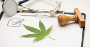 Medical-themed image featuring a green cannabis leaf placed on a white paper, symbolizing Nebraska medical cannabis. A stethoscope with dark blue tubing and a silver chest piece is partially visible in the upper left corner. A gray prescription icon with an 'Rx' and pencil is on the left side of the paper. To the right of the cannabis leaf, a wooden stamp lies on its side, suggesting official approval or certification. The clean white background and bright lighting emphasize the connection between cannabis and medical regulation
