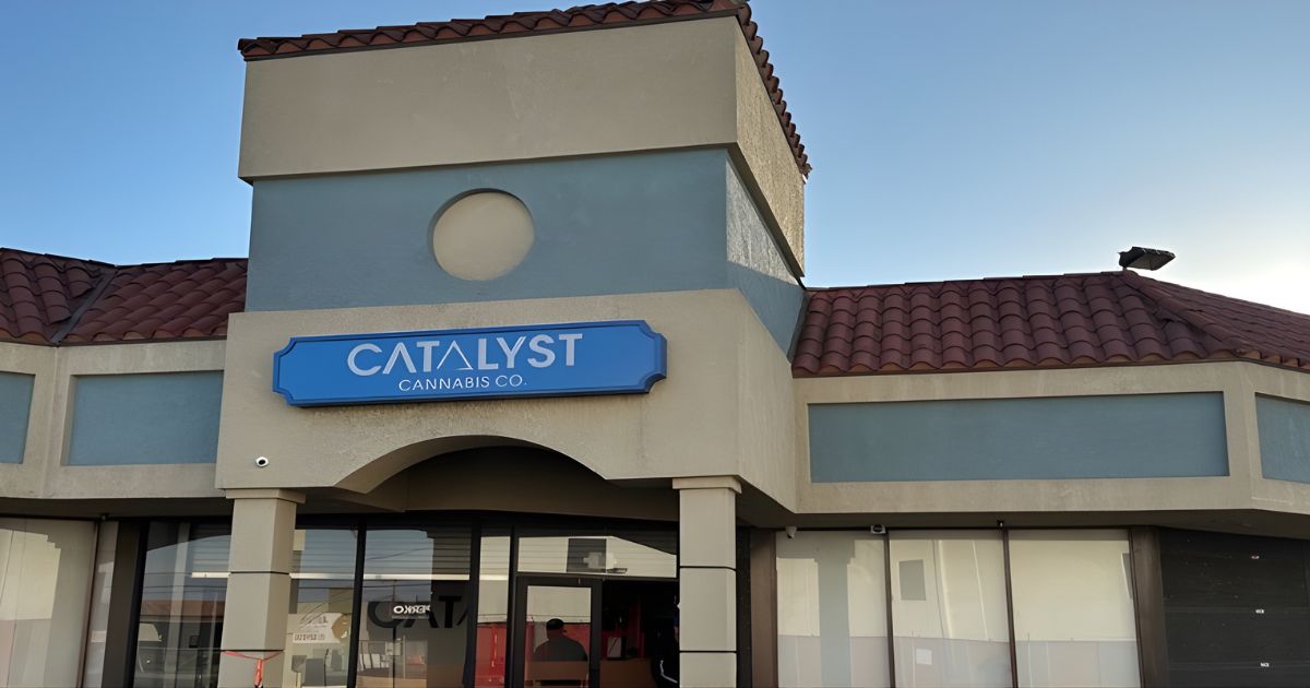 Exterior of the Catalyst Cannabis Co. building in Hawthorne, featuring Mediterranean-style architecture with light stucco walls and a red-tiled roof. A prominent blue sign above the glass-door entrance reads 'CATALYST' in bold white letters, with 'CANNABIS CO.' below it. The entrance is recessed under an archway, and a security camera is mounted on the beige wall. The building sits under a clear blue sky, reflecting a welcoming retail environment.