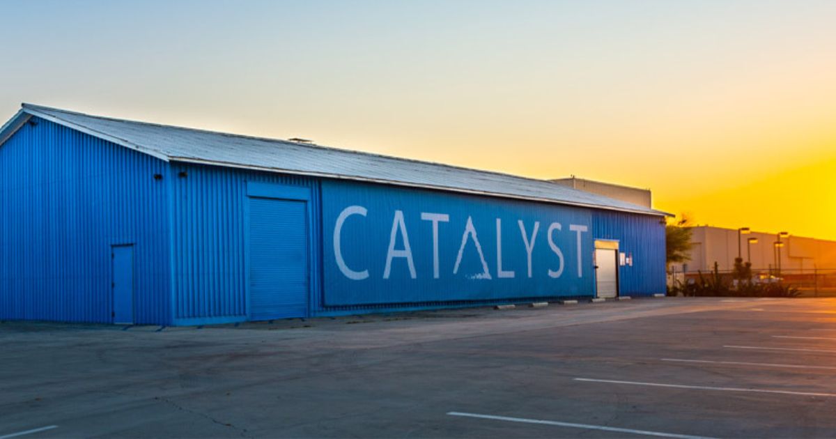 Exterior of the vibrant blue Catalyst Cannabis building on Cherry Ave, featuring bold white 'CATALYST' lettering with stylized triangular 'A's across its corrugated metal facade. The building includes a small door, a large roll-up door, and a reflective entrance, all fronted by a dark grey parking lot with white markings. The background showcases industrial buildings, light poles, and sparse vegetation. A dramatic sky transitions from light blue to fiery orange and yellow, suggesting sunrise or sunset. Keywords: Catalyst cannabis Cherry Ave