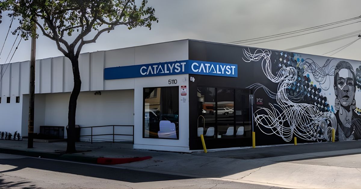 Corner view of the Catalyst Cannabis building in Lynwood, featuring a white facade on the left with the address '5110' and a bold blue strip displaying 'CATALYST' in white text. The right side showcases a dark wall adorned with a vibrant mural, including abstract patterns and a stylized human face. Large glass windows reflect the street, while yellow bollards line the sidewalk. A tree with sparse green leaves and a utility pole stand nearby, under an overcast sky. Keywords: Catalyst Cannabis Lynwood
