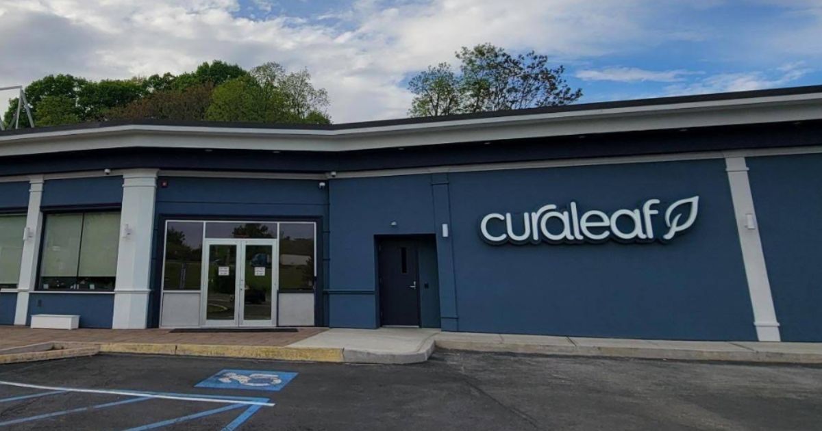 Exterior of the Curaleaf Hudson Valley retail location in Newburgh, New York. The single-story building features a modern blue-grey facade with white trim, a prominent 'curaleaf' logo with a leaf symbol, and large windows. A handicap parking space is visible in the asphalt lot, with green trees and a partly cloudy sky in the background. Keywords: Curaleaf Hudson Valley