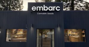Exterior of Embarc South Lake Tahoe, a modern cannabis retail store with a dark vertically paneled facade. The illuminated white 'embarc' sign is prominently displayed above the entrance, with 'Cannabis Goods' written below it. The central glass double doors are flanked by large display windows showcasing warm, inviting interiors with wooden counters and products. Wall-mounted lights enhance the sleek, contemporary design, while tall trees in the background suggest a natural setting. Keywords: Embarc South Lake Tahoe