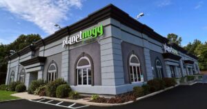 Exterior view of the Planet Nugg building in Farmingdale, New York, featuring a modern design with light and dark gray tones and distinctive arched windows. The 'planetnugg' logo, with 'planet' in white and 'nugg' in green, is prominently displayed on the building. Surrounded by well-maintained landscaping, a parking lot, and a clear blue sky, the setting highlights the suburban location. This signifies the Planet Nugg Farmingdale retail spotlight