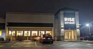Nighttime view of the RISE Dispensary in Henrietta, New York. The modern building features a prominent illuminated sign reading 'RISE DISPENSARY' above the stone-clad main entrance. Large windows reveal a brightly lit interior, while a black sedan is parked in the foreground of the paved parking lot with visible handicap markings. Keywords: Rise Dispensary Henrietta