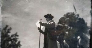 Grainy black-and-white historical photograph of a man, identified as Robert Levers, wearing a tricorn hat and holding a document in one hand and a walking stick in the other. He appears to be reading or addressing an audience outdoors, with a bright overcast sky and dark foliage in the background. Keywords: Robert Levers, Declaration of Independence, Easton Pennsylvania