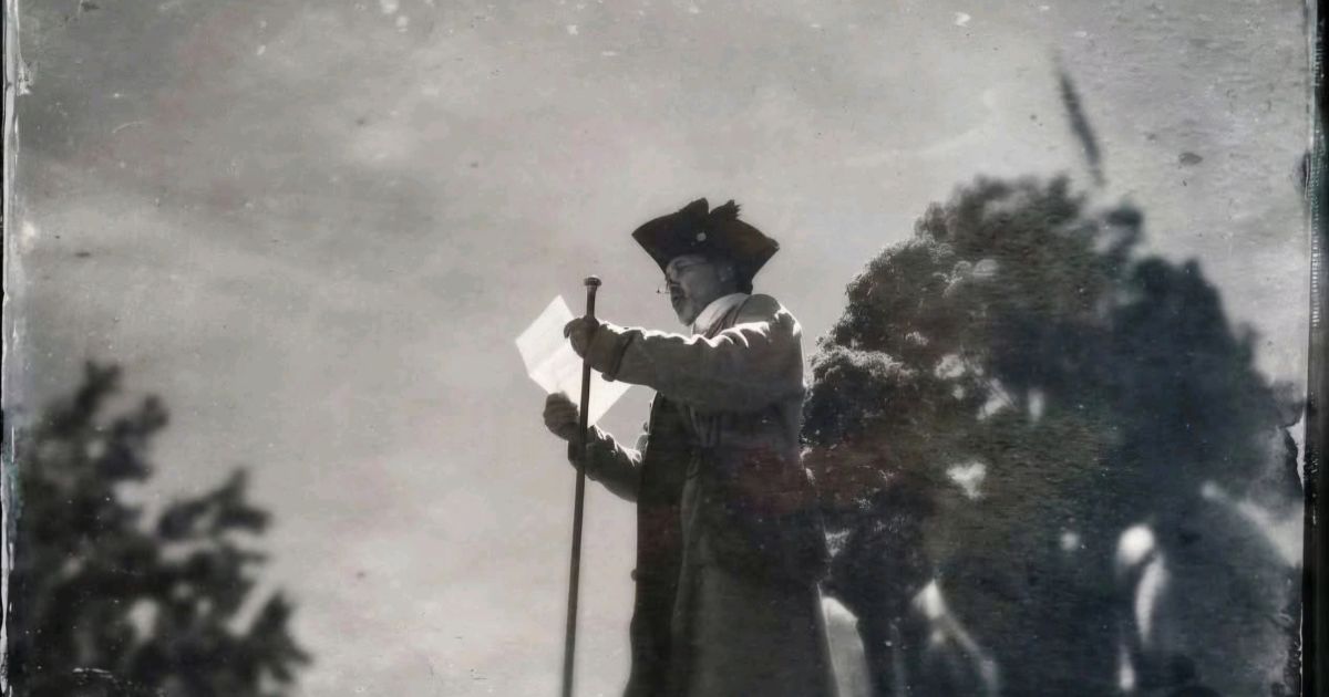 Grainy black-and-white historical photograph of a man, identified as Robert Levers, wearing a tricorn hat and holding a document in one hand and a walking stick in the other. He appears to be reading or addressing an audience outdoors, with a bright overcast sky and dark foliage in the background. Keywords: Robert Levers, Declaration of Independence, Easton Pennsylvania