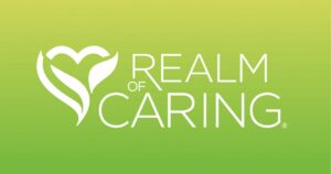 Logo for Realm of Caring displayed on a gradient green background transitioning from light yellow-green at the top to dark green at the bottom. On the left, a white stylized heart or plant-like emblem with leaf shapes is featured. To the right, the text 'REALM OF CARING' appears in white uppercase sans-serif font, with 'CARING' being the largest word. A registered trademark symbol (®) is positioned next to 'CARING.