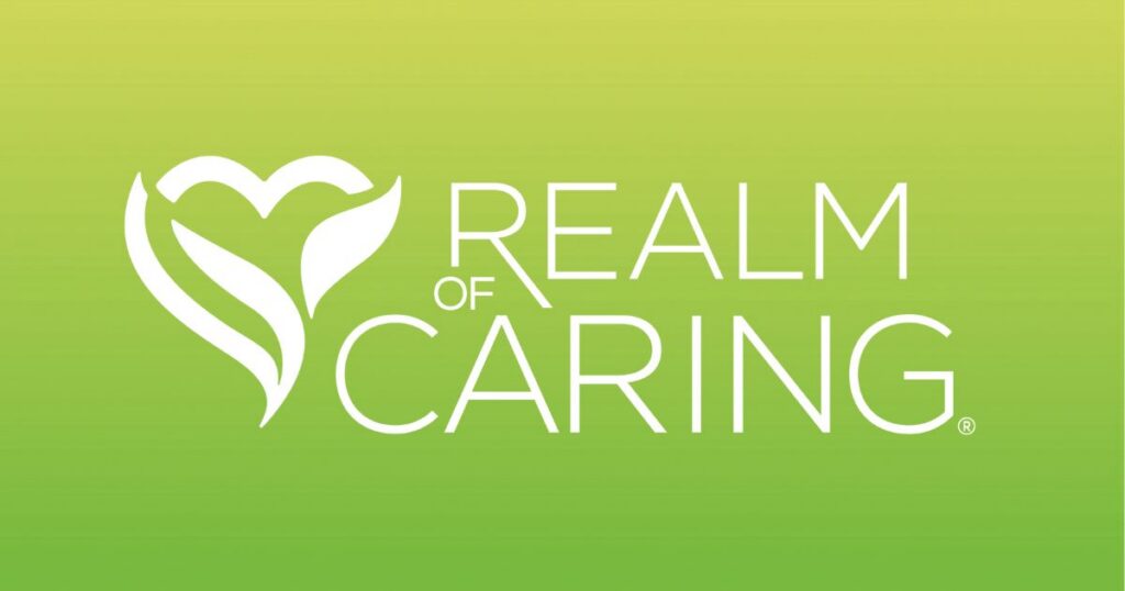 Logo for Realm of Caring displayed on a gradient green background transitioning from light yellow-green at the top to dark green at the bottom. On the left, a white stylized heart or plant-like emblem with leaf shapes is featured. To the right, the text 'REALM OF CARING' appears in white uppercase sans-serif font, with 'CARING' being the largest word. A registered trademark symbol (®) is positioned next to 'CARING.