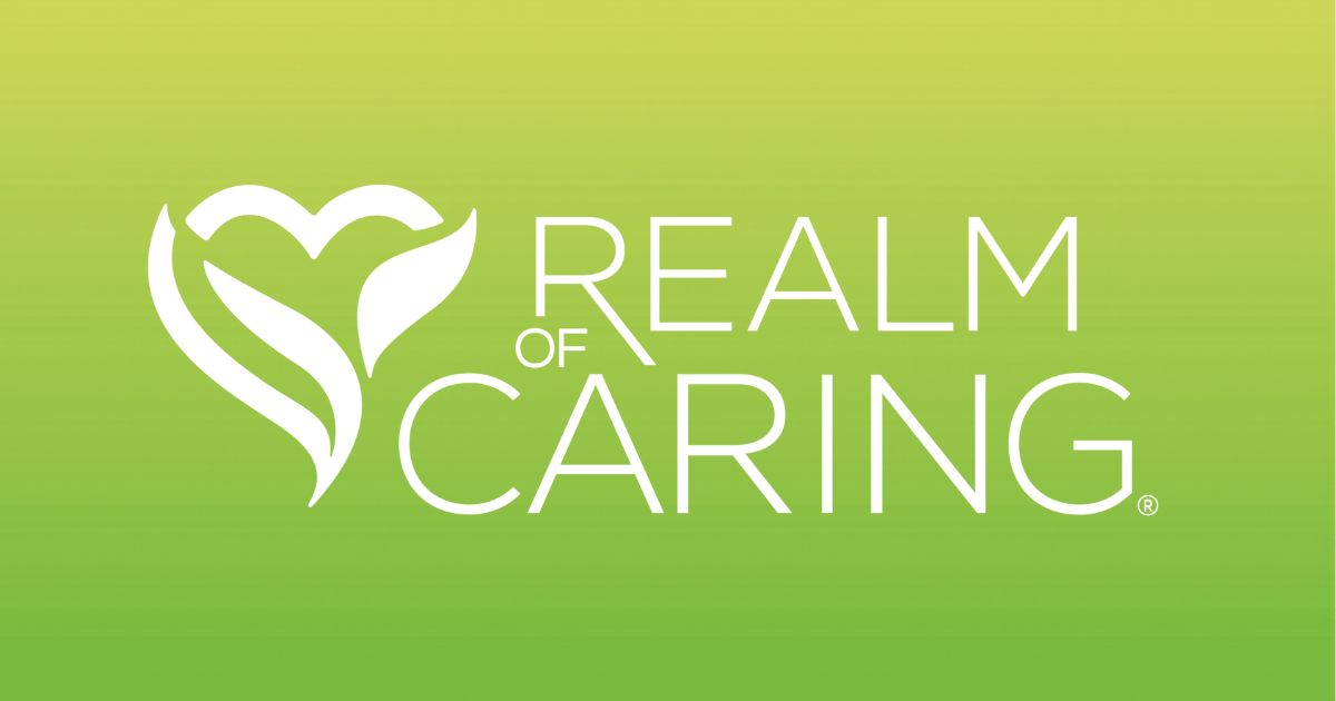 Logo for Realm of Caring displayed on a gradient green background transitioning from light yellow-green at the top to dark green at the bottom. On the left, a white stylized heart or plant-like emblem with leaf shapes is featured. To the right, the text 'REALM OF CARING' appears in white uppercase sans-serif font, with 'CARING' being the largest word. A registered trademark symbol (®) is positioned next to 'CARING.