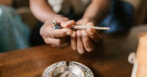 Close-up of dark-skinned hands holding a hand-rolled joint and a light blue disposable lighter, positioned above a round, ornate silver ashtray on a warm wooden surface. The joint appears ready to be lit, with loose material visible at the end. The blurred background suggests an indoor setting with hints of teal fabric and light-colored furniture. Keywords: Virginia House Bill 942