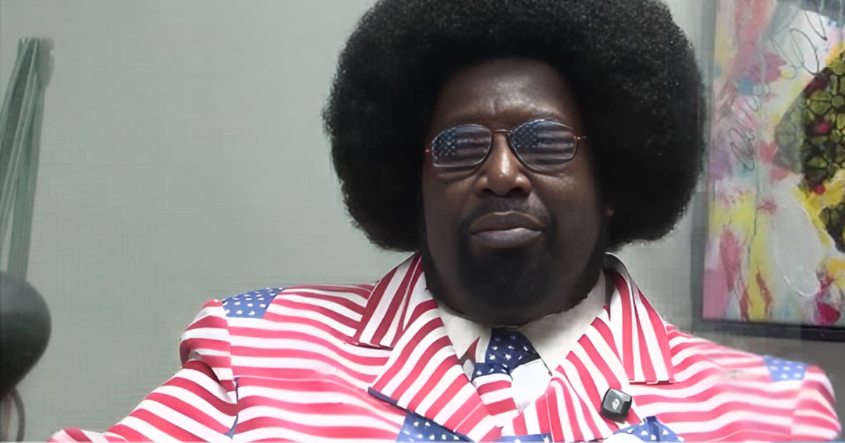 Medium close-up of Afroman, wearing sunglasses reflecting the American flag and a suit patterned with the flag's design. He looks directly at the viewer with a neutral expression. The background features a plain wall on the left and a colorful abstract painting on the right, symbolizing the Afroman case and themes of patriotism and justice