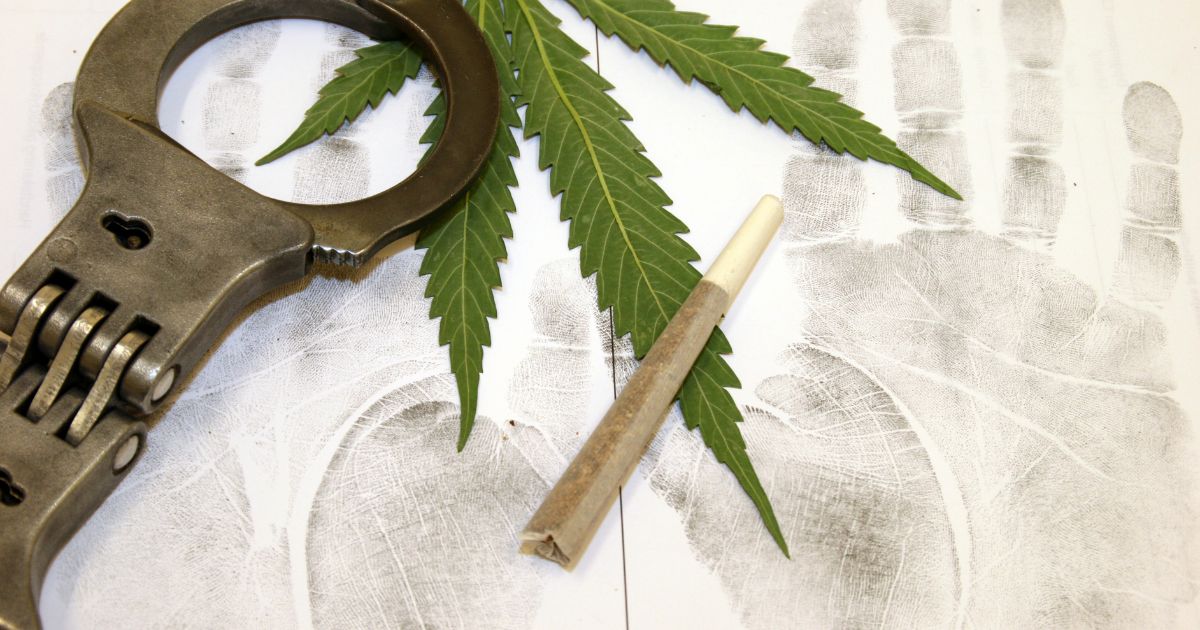 Conceptual image representing cannabis enforcement in the US, featuring a vibrant green cannabis leaf, a rolled joint, and metallic handcuffs placed on a white background covered with black forensic palm and fingerprints, symbolizing the intersection of marijuana use and law enforcement