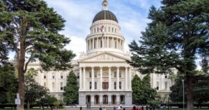 California State Capitol building in Sacramento, featuring a grand Neoclassical design with a white dome, Corinthian columns, and intricate pediment sculptures. Framed by tall evergreen trees, the scene includes a bicycle lane sign in the foreground and people scattered across the plaza, symbolizing the civic context of AB 2532