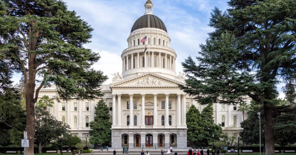 California State Capitol building in Sacramento, featuring a grand Neoclassical design with a white dome, Corinthian columns, and intricate pediment sculptures. Framed by tall evergreen trees, the scene includes a bicycle lane sign in the foreground and people scattered across the plaza, symbolizing the civic context of AB 2532