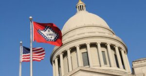 Low-angle view of the Arkansas State Capitol dome with the Arkansas state flag and U.S. flag waving on silver poles in the foreground. The Arkansas flag, featuring a red field and white diamond, symbolizes state pride, set against a clear blue sky, referencing Arkansas hemp legislation