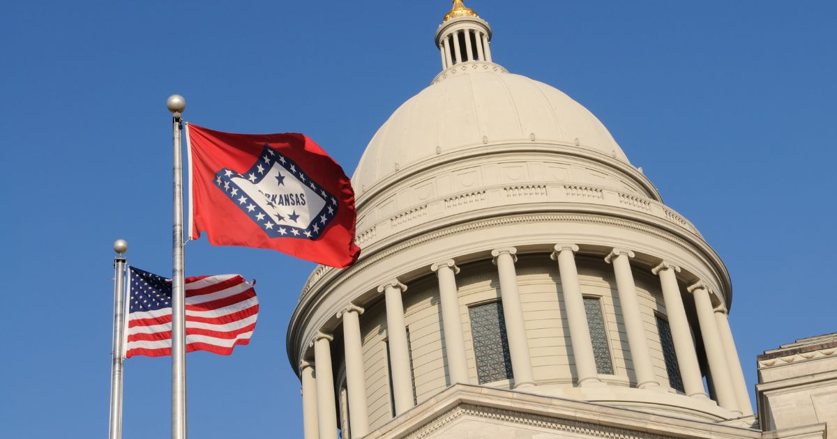 Low-angle view of the Arkansas State Capitol dome with the Arkansas state flag and U.S. flag waving on silver poles in the foreground. The Arkansas flag, featuring a red field and white diamond, symbolizes state pride, set against a clear blue sky, referencing Arkansas hemp legislation