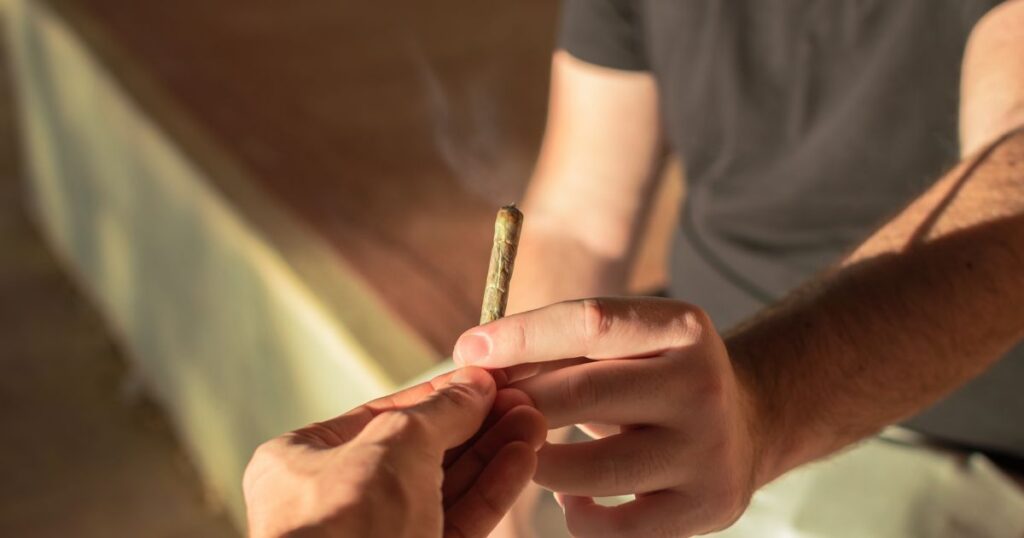 Close-up of two hands passing a lit hand-rolled cannabis joint, with a faint wisp of smoke rising. The scene is warmly lit by golden hour sunlight, creating a relaxed and intimate atmosphere, symbolizing themes related to cannabis use among youth