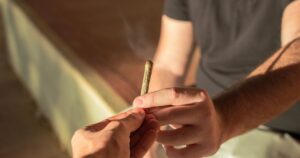 Close-up of two hands passing a lit hand-rolled cannabis joint, with a faint wisp of smoke rising. The scene is warmly lit by golden hour sunlight, creating a relaxed and intimate atmosphere, symbolizing themes related to cannabis use among youth