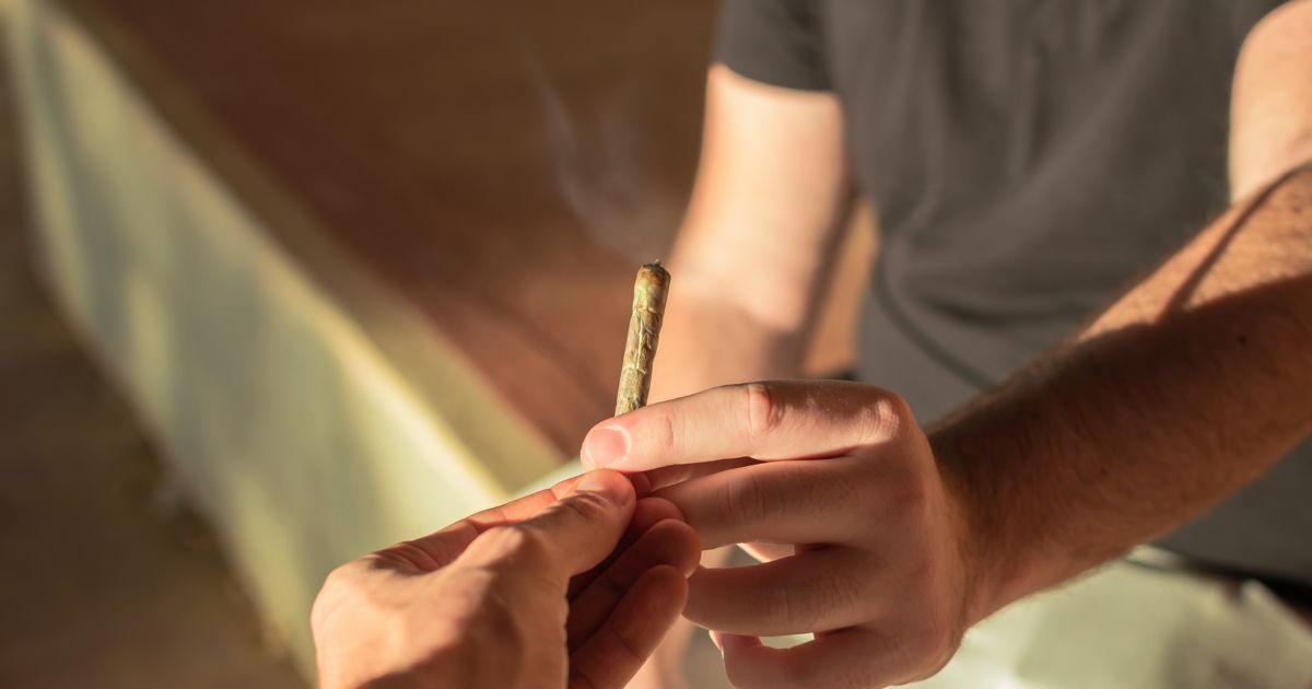 Close-up of two hands passing a lit hand-rolled cannabis joint, with a faint wisp of smoke rising. The scene is warmly lit by golden hour sunlight, creating a relaxed and intimate atmosphere, symbolizing themes related to cannabis use among youth