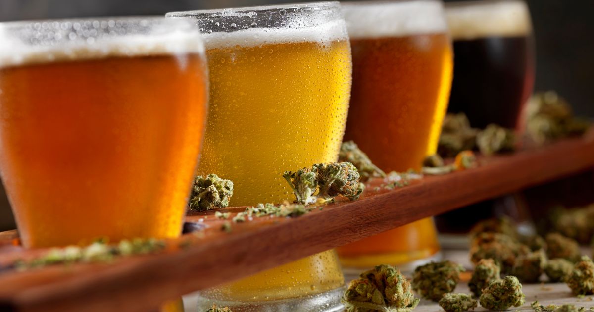 Close-up of a wooden flight board holding four craft beer glasses, ranging from golden to dark amber, with cannabis buds scattered around the glasses and on the board. The shallow depth of field highlights the golden beer and nearby cannabis, symbolizing the DC THC drink partnership with local breweries and distilleries