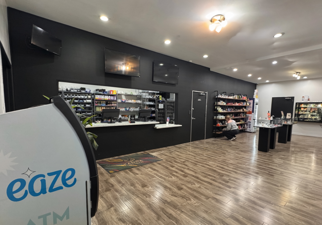 Interior of an Eaze cannabis dispensary featuring a modern design with a matte black feature wall, white service counter, and wood-textured flooring. The foreground includes an Eaze-branded ATM, while the middle ground showcases glass display cases, shelving with cannabis products, and a staff member organizing items. Bright lighting and clean aesthetics create a professional retail environment
