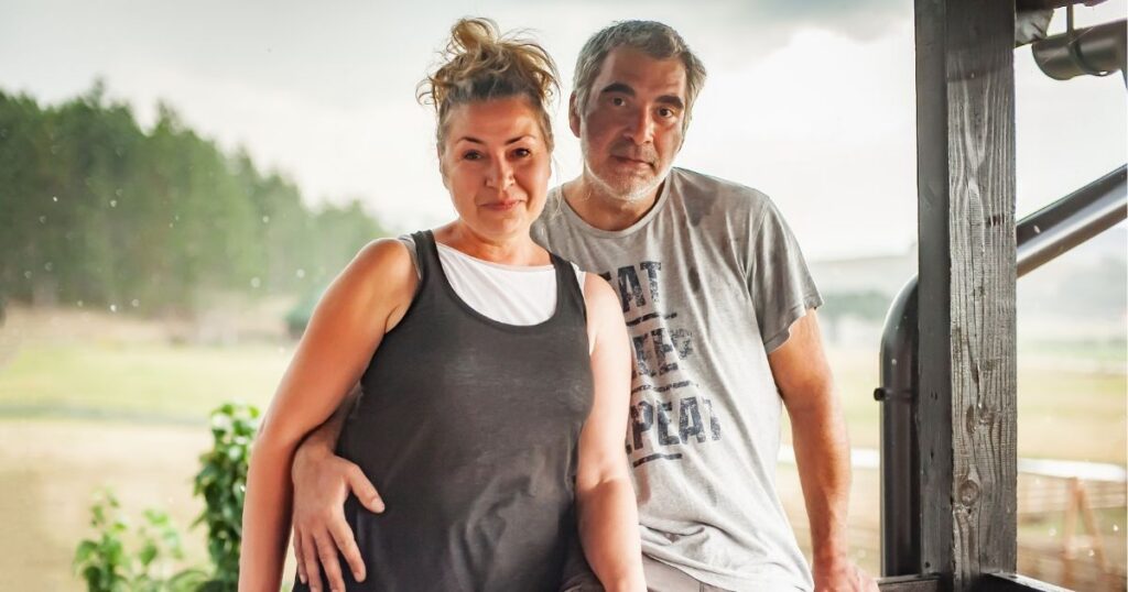 A smiling couple stands on a porch during light rain, with the man wearing a gray 'EAT SLEEP REPEAT' t-shirt and the woman in casual attire. The background features a grassy field and trees, symbolizing a relaxed moment tied to cannabis legalization discussions