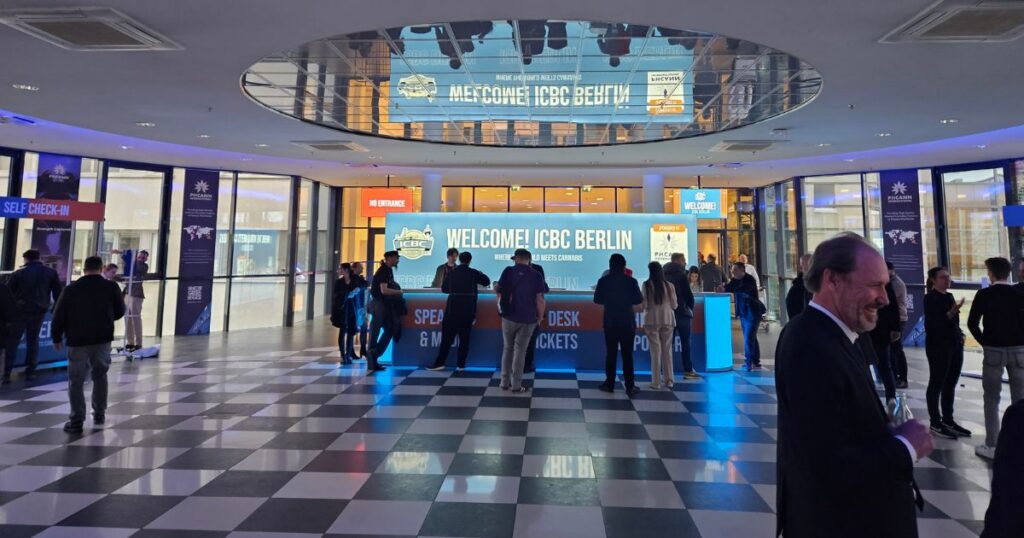 Lobby of the ICBC Berlin conference featuring a brightly lit blue welcome desk with 'Welcome! ICBC Berlin' and 'Where World Meets Cannabis' signage. The modern space includes black and white checkered flooring, a mirrored ceiling, and glass walls with banners for sponsor PHCANN International. Attendees in professional attire gather around the desk, emphasizing the international cannabis business summit