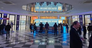 Lobby of the ICBC Berlin conference featuring a brightly lit blue welcome desk with 'Welcome! ICBC Berlin' and 'Where World Meets Cannabis' signage. The modern space includes black and white checkered flooring, a mirrored ceiling, and glass walls with banners for sponsor PHCANN International. Attendees in professional attire gather around the desk, emphasizing the international cannabis business summit