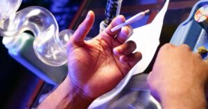Close-up of a hand holding a hand-rolled cannabis joint, surrounded by paraphernalia including a glass bong with smoke, a blue rolling tray, a cannabis bud, and a grinder. The scene is lit with dramatic blue and purple lighting, symbolizing the debate over Maine's cannabis consumption lounge bill