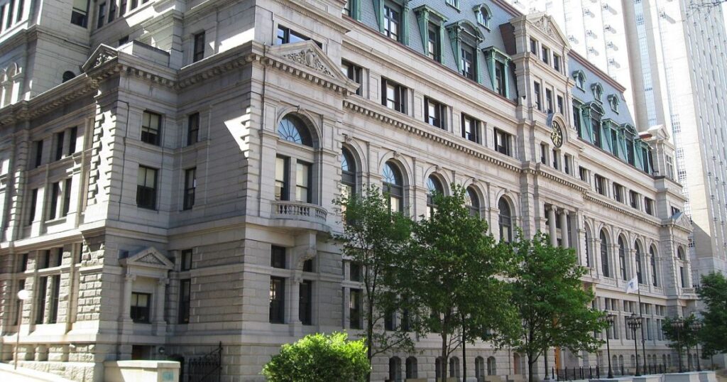 Low-angle view of a historic Beaux-Arts style building, likely a courthouse in Massachusetts, with arched windows, a mansard roof, and a central clock. Green trees partially obscure the facade, while a modern skyscraper rises in the background. The scene symbolizes the Massachusetts cannabis operators' lawsuit challenging a ballot initiative