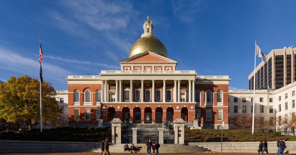 Daytime view of the Massachusetts State House in Boston, featuring its iconic golden dome, red brick façade, and white Corinthian columns. The foreground includes a stone plaza, stairs, and autumn trees with yellow foliage, symbolizing the state's modernization efforts, including cannabis consumption lounges