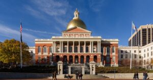 Daytime view of the Massachusetts State House in Boston, featuring its iconic golden dome, red brick façade, and white Corinthian columns. The foreground includes a stone plaza, stairs, and autumn trees with yellow foliage, symbolizing the state's modernization efforts, including cannabis consumption lounges