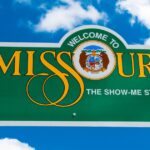 Missouri welcome sign featuring 'WELCOME TO MISSOURI' in gold serif font with the state seal replacing the 'O' and the tagline 'THE SHOW-ME STATE' below. Set against a bright blue sky with fluffy clouds, symbolizing Missouri cannabis operators lawsuit against Good Day Farm