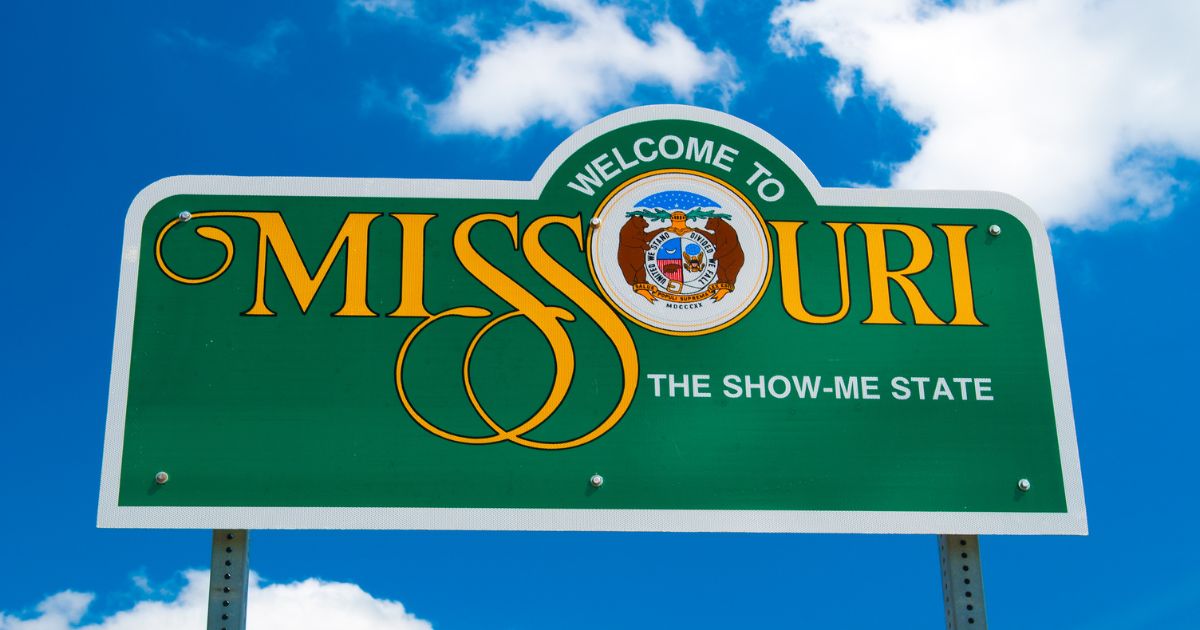 Missouri welcome sign featuring 'WELCOME TO MISSOURI' in gold serif font with the state seal replacing the 'O' and the tagline 'THE SHOW-ME STATE' below. Set against a bright blue sky with fluffy clouds, symbolizing Missouri cannabis operators lawsuit against Good Day Farm