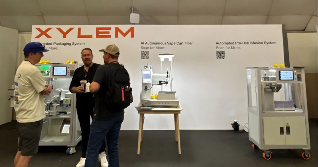 Trade show booth for Xylem Robotics at the 2026 Hall of Flowers, showcasing automated cannabis manufacturing equipment. Three machines are displayed: a packaging system, an AI-driven vape cartridge filler, and a pre-roll infusion system. Attendees interact near the machines, which feature digital interfaces and QR codes for more information. The booth is branded with large red 'XYLEM' lettering, emphasizing innovation in cannabis production technology
