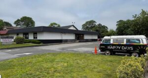Exterior of Happy Days Dispensary in Farmingdale, New York, featuring a single-story white building with a black roof, a monument sign displaying the business name, and a retro-style Volkswagen van with tie-dye accents and 'Happy Days Dispensary' branding parked in the lot. The scene is framed by green landscaping and an overcast sky