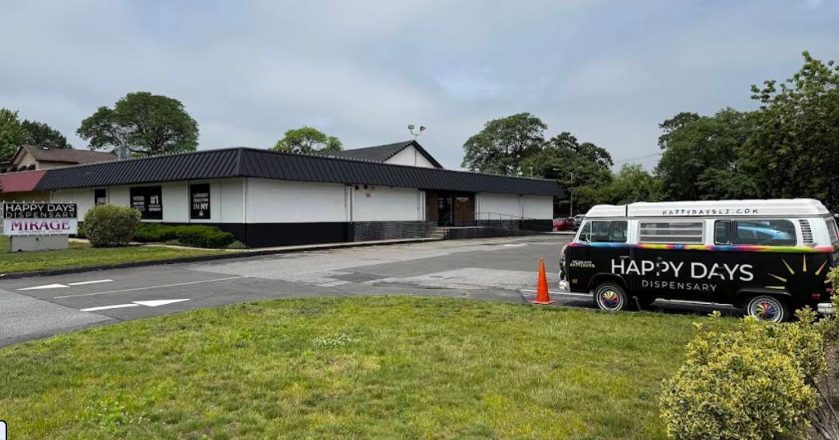 Exterior of Happy Days Dispensary in Farmingdale, New York, featuring a single-story white building with a black roof, a monument sign displaying the business name, and a retro-style Volkswagen van with tie-dye accents and 'Happy Days Dispensary' branding parked in the lot. The scene is framed by green landscaping and an overcast sky