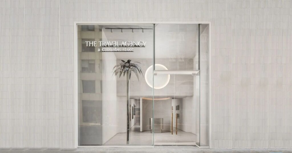 Front view of 'The Travel Agency,' a modern cannabis store on 5th Ave in Manhattan, New York. The storefront features a large glass facade framed by light, vertically striped tiles. Inside, a metallic palm tree sculpture and a glowing circular light create a sleek, upscale ambiance. The store's name, 'The Travel Agency,' and tagline, 'a cannabis store,' are prominently displayed, reflecting its contemporary branding
