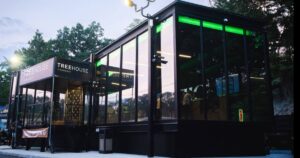 Modern cannabis dispensary 'Treehouse Cannabis' in Nyack, NY, featuring a sleek black metal and glass structure with vibrant green LED interior lighting. The storefront includes large glass windows, a raised entrance with 'NOW OPENING' banners, and a backdrop of dense trees under an overcast sky, highlighting its contemporary design