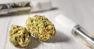 Close-up of two dried cannabis buds with green and amber tones, placed alongside a vaporizer cartridge featuring a white mouthpiece and golden oil reservoir. The image contrasts traditional cannabis flower with modern cannabis vape options, set on a bright, textured white surface