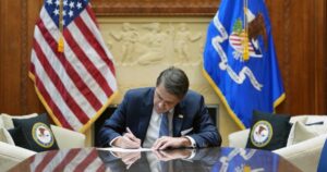 Formal scene of a U.S. government official in a navy suit signing a document at a polished wooden table, flanked by the American flag and the Department of Justice flag. The setting includes DOJ-branded pillows and a decorative wall relief, symbolizing the official action to reschedule marijuana