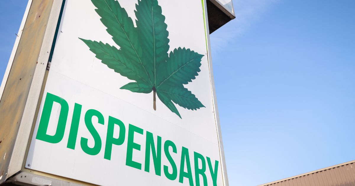 Low-angle view of a cannabis dispensary sign in Virginia featuring a bold green cannabis leaf graphic above the word 'DISPENSARY' in uppercase green letters. The sign is set against a clear blue sky with part of a brown corrugated building visible in the corner, symbolizing the Virginia cannabis market